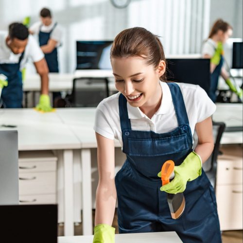 cheerful cleaner in overalls cleaning office desk with rag